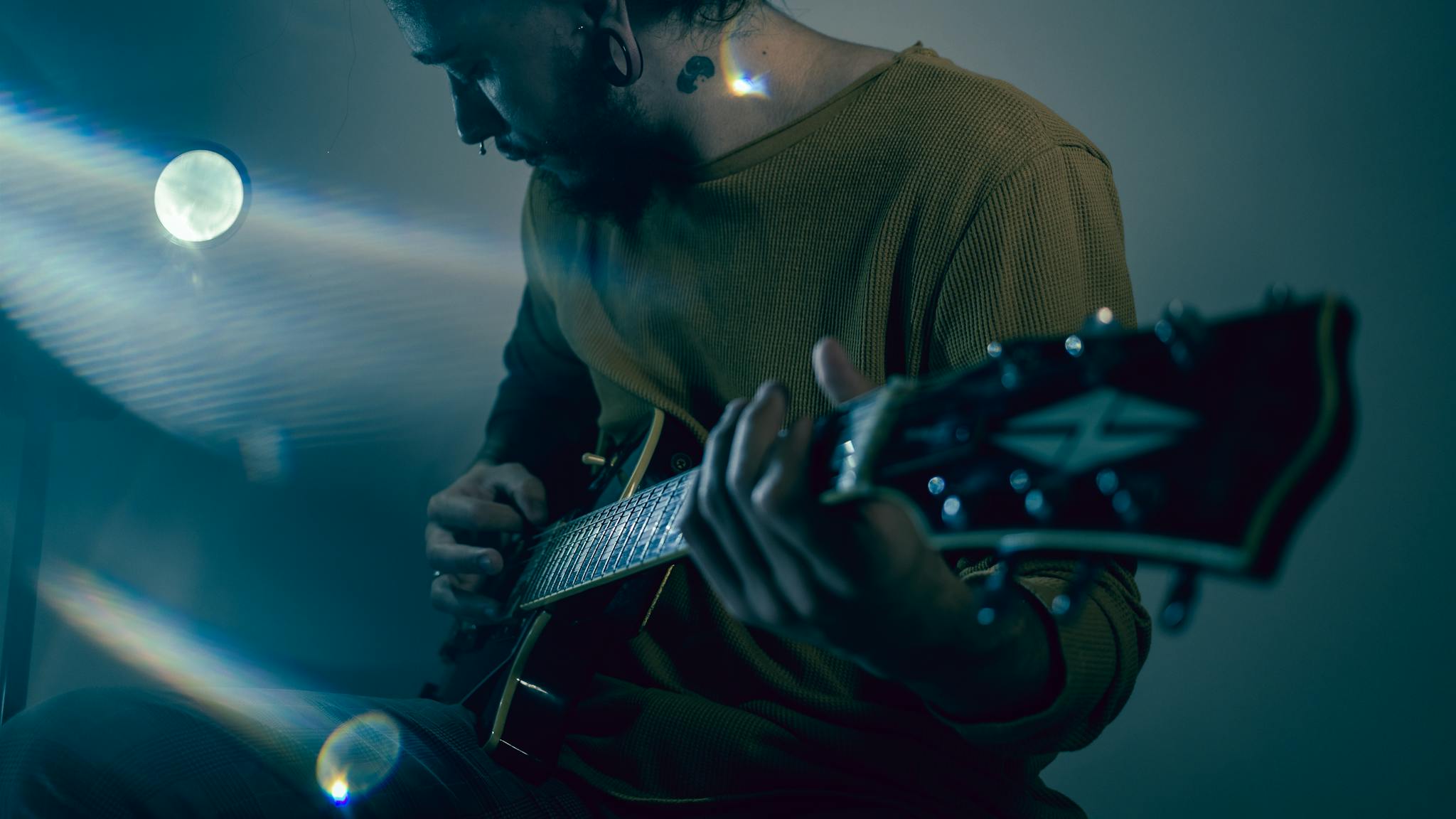 A man in a sweater plays an electric guitar against a moody background with artistic lighting.