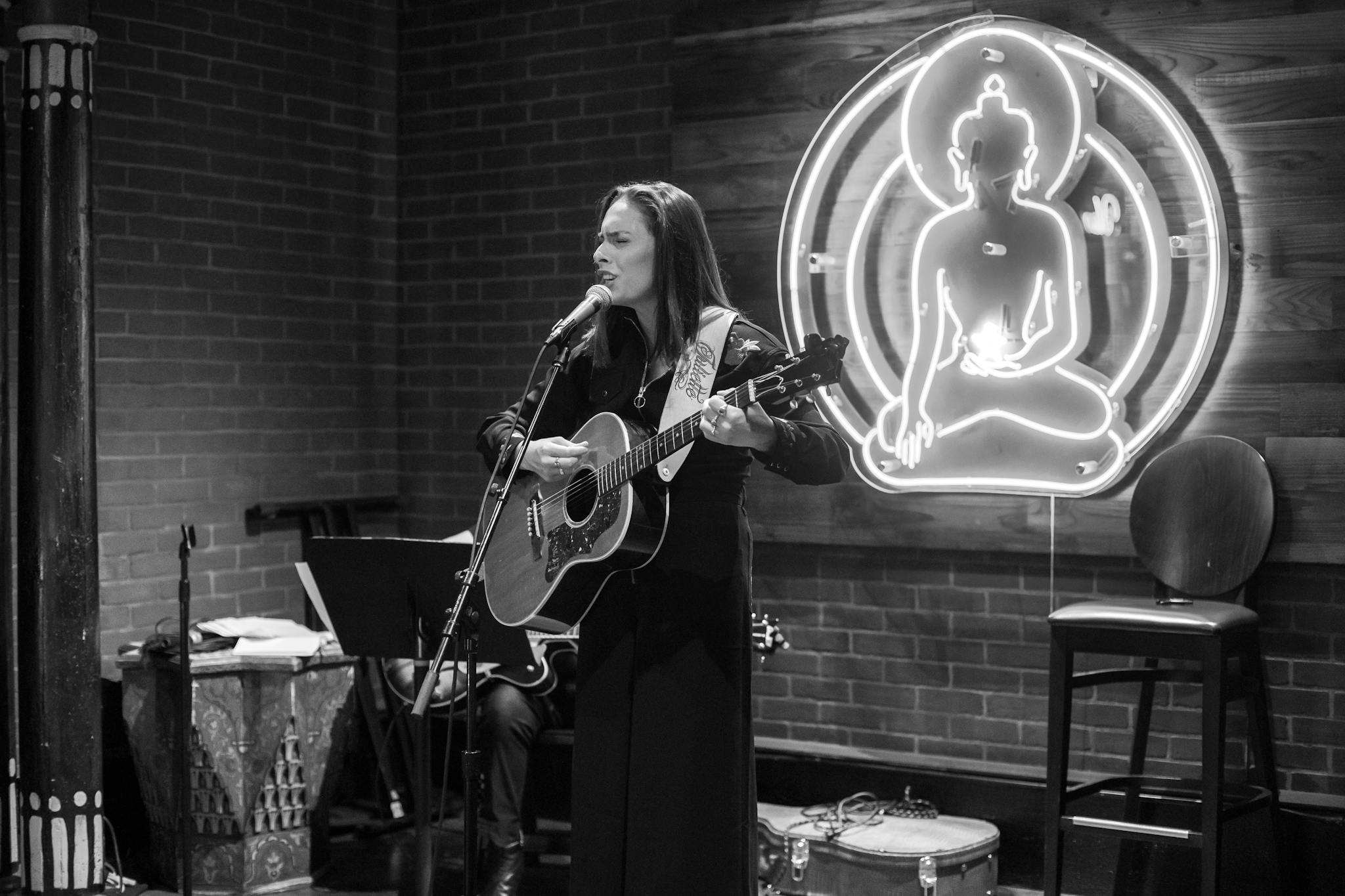 A woman performs with a guitar in a cozy, brick-wall venue featuring a glowing Buddha sign.