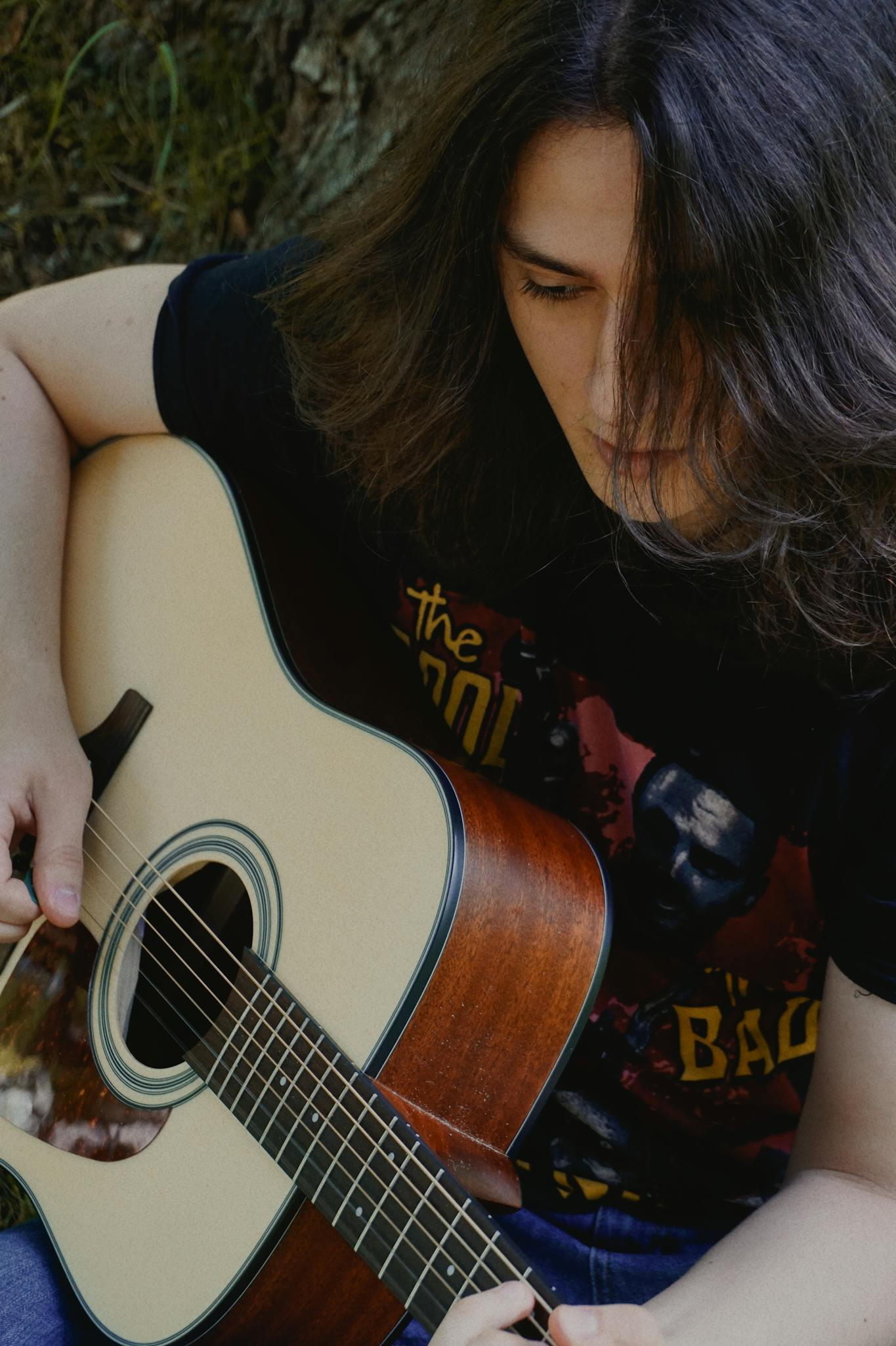 A young adult with long hair playing an acoustic guitar in a serene outdoor setting.
